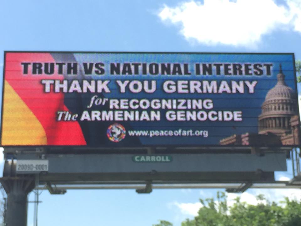 Titled "Truth Vs. National Interest," this billboard illustrates, on the left side, the German flag and, on the opposite side, the dome of the U.S. Capitol building, where the United States Senate and House of Representatives come together to debate and discuss national and political issues.